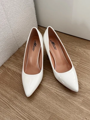 Vita klackskor Studenten - Snygga vita pumps från utomlands  i glansigt lackmaterial. Skorna har spetsig tå och en stabil blockklack som ger både höjd och komfort. Perfekta för att lyfta vilken outfit som helst med sin stilrena och moderna look.perfekta till STUDENTEN eller annan högtid nu på sommarn/fester.