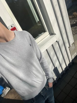Ljusgrå tröja från Polo Ralph Lauren - Snygg ljusgrå tröja från Polo Ralph Lauren med klassisk rund halsringning och diskret logga broderad på bröstet. Tröjan har långa ärmar och ribbade muddar vid ärmslut och nederkant. Perfekt till jeans för en clean look.