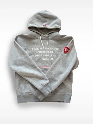 Grå hoodie från Peak Performance - Snygg grå hoodie från Peak Performance med vit text på bröstet och röd logga på ärmen.  Skick 10/10