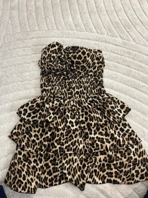 Axelbandslös leopardklänning volanger - Säljer en axelbandslös klänning med leopardmönster och flera lager volanger. Klänningen har smock i midjan och en söt rosett framtill. Materialet känns lätt och luftigt, perfekt för sommarens alla tillfällen. Djurmönstret ger en trendig och cool vibe.