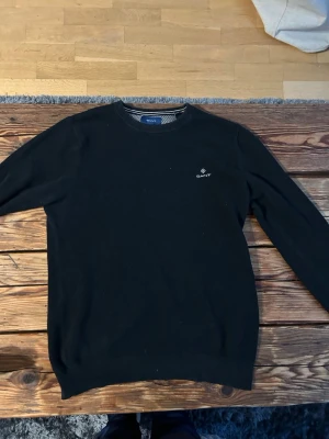 Stickad gant tröja - Klassisk svart tröja från GANT med rund halsringning och diskret broderad logga på bröstet. Tröjan har ribbade muddar vid ärmslut och nederkant samt ett snyggt mönstrat innerfoder vid halsen. Perfekt för en stilren look. Pris kan diskuteras