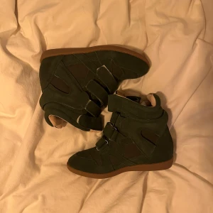 Isabel Marant Bekett - 38 - Säljer ett par mörkgröna sneakers med kilklack och kardborreband. Skorna har en cool design med paneler i mocka och textil samt gummisula i brunt. Perfekta för dig som vill ha både stil och höjd i vardagen.