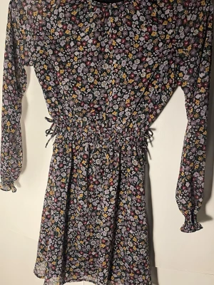 Blommig kortklänning med långa ärmar - Storlek 146 men passar mig som bär 34/36!!! Den är stretchig. Supersöt kortklänning med svart botten och färgglatt blommönster i gult, lila, vitt och rött. Endast andvänd fåtal gånger. Klänningen har långa ärmar med resår vid handlederna och markerad midja med snörning i sidorna. Perfekt för dig som gillar romantisk och lekfull stil.