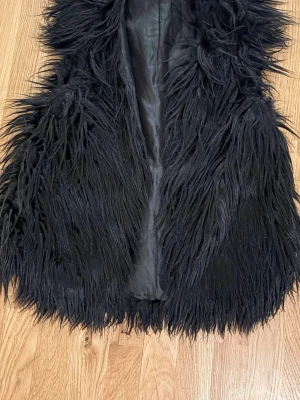 Svart pälsväst från Gina Tricot - Säljer en svart pälsväst från Gina Tricot med lång, fluffig fuskpäls och slät insida. Västen har öppen front och ger en riktigt cool och trendig look. Perfekt att slänga över en hoodie eller tröja för extra edge.