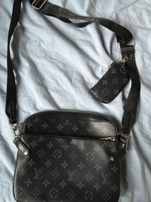  svart axelväska monogram - Snygg svart axelväska med klassiskt monogrammönster i grått. Väskan har justerbar axelrem och kommer med en matchande liten pouch. Tillverkad i slitstarkt konstläder med silverfärgade detaljer och dragkedja. Perfekt för dig som gillar lyxig streetstyle. Kan diskutera pris 