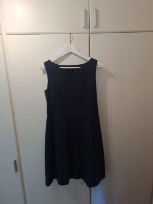 Marinblå ärmlös klänning - Klassisk marinblå klänning med ärmlös design och enkel, stilren siluett. Klänningen har en liten v-ringning framtill och är knälång med minimalistisk och tidlös stil. Bomull med polyesterfoder ichbdragkedja mitt bak. Observera att jag gjort ett hål i fodret vid ringningen. I övrigt är klänningen i bra skick.