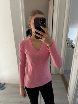 Rosa stickad tröja med V-ringning - Snygg rosa stickad tröja med V-ringning och långa ärmar. Tröjan har en liten broderad logga på bröstet och är figurnära i modellen. 