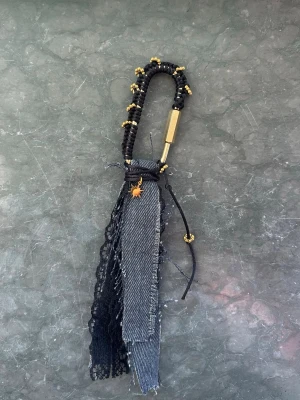 väsksmycke i denim - Unikt väsksmycke med svarta och guldiga pärlor, denimtyg och spetsdetaljer. Perfekt att hänga på väskan för en personlig touch. Har även en liten guldfärgad berlock och karbinhake för enkel fastsättning.