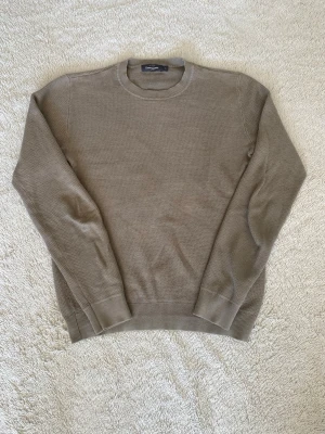Beige stickad tröja från Dressman - Snygg beige stickad tröja från Dressman med rund halsringning och ribbade muddar vid ärmslut och nederkant. Tröjan har en enkel och stilren design som passar perfekt till många olika outfits. Materialet känns mjukt och bekvämt. Färgen är brun/mörkgrön aktig.