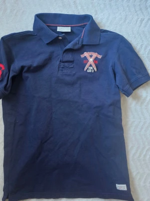 Marinblå pikétröja Peak Performance - Snygg marinblå pikétröja från Peak Performance med broderad logga och detaljer i rött och vitt på bröstet. Klassisk krage med knappslå och korta ärmar. Perfekt till jeans eller shorts för en sportig look.