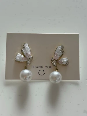 Fjärilsörhängen med pärldetaljer - Säljer ett par unika örhängen i form av fjärilar med skimrande vita och rosa stenar, guldfärgade kanter och stora hängande pärlor. Örhängena har en elegant och lekfull design som verkligen sticker ut.