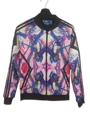 Adidas Originals x The Farm Company "Florera" Track Top, S - Denna Adidas-jacka har en livfull design med fåglar och blommor i nyanser av lila, rosa och blått. Jackan har en hel dragkedja framtill och ribbade muddar i ärmslut och nederkant. Adidas logotypen är placerad på vänster bröst och en större version på ryggen. De klassiska tre ränderna löper längs ärmarna i svart, vilket ger en sportig kontrast till det färgglada mönstret. Armhåla till armhåla: 46cm Axelbredd: 41cm Långt: 61cm Armlängden från armhåla: 52cm