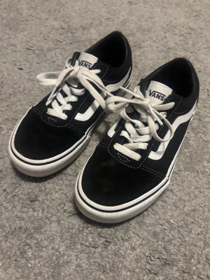 Svarta Vans Old Skool sneakers - Säljer ett par klassiska Vans Old Skool sneakers i svart och vitt med vit sula och vita snören. Skorna har den ikoniska sidoremsan och är tillverkade i canvas och mocka. Perfekta för dig som gillar streetstyle och vill ha en tidlös look. Använda max 3 ggr. 