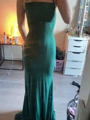 Bal kläning - Säljer en elegant grön långklänning med axelbandslös design och figurnära passform. Klänningen har en släp-liknande nederdel och är tillverkad i ett mjukt, följsamt tyg som ger en snygg siluett. Perfekt för dig som vill sticka ut med en stilren look.