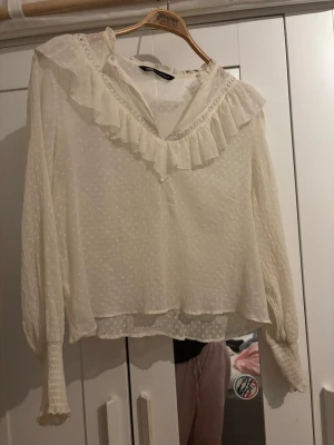 Vit blus med volang från Zara - Söt vit blus från Zara med volangdetaljer längs V-ringningen och diskret prickigt mönster i tyget. Blusen har långa ärmar med smock vid ärmsluten och är gjord i ett tunt, lätt material som ger en romantisk känsla.