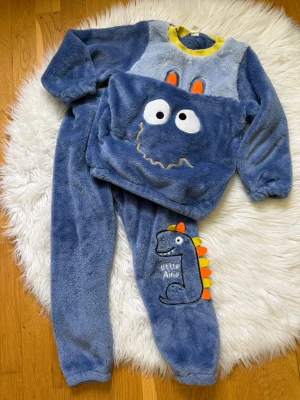Blå fluffig pyjamas med dinosauriemotiv - Supermjuk och mysig blå pyjamas i fleece med dinosauriemotiv. Tröjan har broderade ögon, orange detaljer och gul kant vid halsen. 