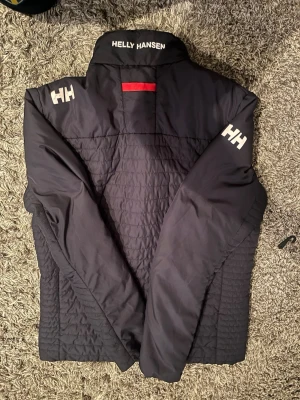 Svart vindjacka från Helly Hansen - Svart vindjacka från Helly Hansen med vita HH-loggor på ärmar och krage. Jackan har ståkrage, röda detaljer vid dragkedjor och quiltade partier på framsidan. Två sidofickor med dragkedja och en bröstficka. Perfekt för blåsiga dagar.
