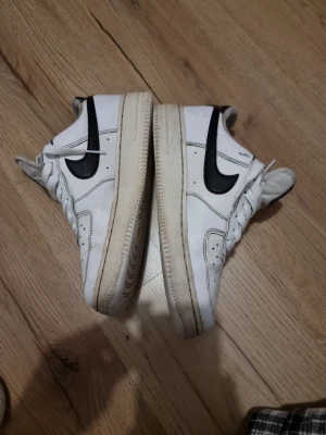 Nike Air Force 1 vita sneakers - Klassiska Nike Air Force 1 sneakers i vitt läder med svart swoosh på sidan. Lågt skaft, perforerad tåbox och vit sula med beige inslag. Skön passform och tidlös design som funkar till det mesta.
