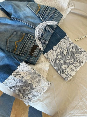 Blå jeans med vit spetsdetalj - Unika blå jeans med utsvängda ben och bred vit spets med blommönster längst ner. Jeansen har dekorativa fickor med gul söm och en cool vintagekänsla. Perfekta för dig som vill sticka ut med en kreativ och trendig look. Egensydda💗💗