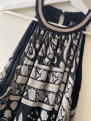 Festklänning/aftonklännig  - Snygg svart långklänning med vitt paisley- och blommönster. Klänningen har omlottmodell med knytband i midjan och är tillverkad i ett tunt, luftigt material som ger ett elegant fall. Perfekt för dig som gillar bohemiska vibes och vill sticka ut.