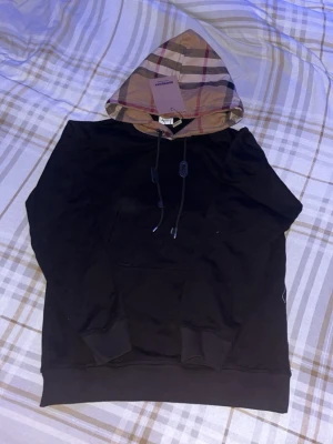 Svart Burberry hoodie med rutmönstrad huva - Säljer en svart hoodie från Burberry med den klassiska beigea rutmönstrade huvan och svarta dragsnören. Hoodien har en stor magficka och ribbade muddar vid ärmar och nederkant. Perfekt för dig som vill ha en stilren men ändå lyxig look.