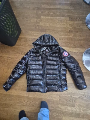 Svart dunjacka från Canada Goose - Säljer en svart dunjacka från Canada Goose med huva och dragkedja framtill. Jackan har en glansig finish och klassisk quiltad design 2 hål i luvan men inget som märks annars är den som ny! Kan lösa bättre bilder 
