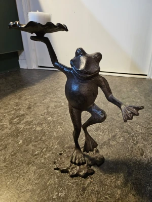 Groda ljushållare i gjutjärn - Unik ljushållare i form av en groda som står på ett ben och håller ett fat för värmeljus. Tillverkad i mörkt gjutjärn med detaljerad design och lekfull pose. Perfekt som dekoration i hemmet för dig som gillar udda inredningsdetaljer. Ca 30 cm hög.