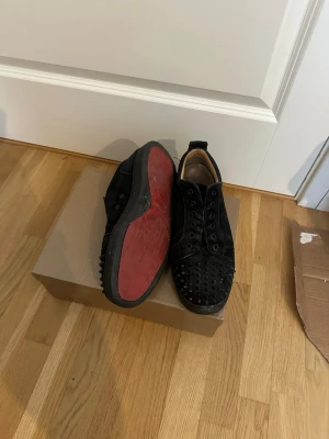 Svarta sneakers med nitar, Louboutin - Svarta låga sneakers från Christian Louboutin med klassiska röda sulor och svarta nitar över hela ovandelen. Skorna har snörning och är tillverkade i mocka för en lyxig känsla. Perfekta för dig som vill sticka ut med en edgy och exklusiv stil.