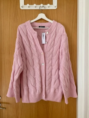 Ljusrosa kabelstickad kofta Gina Tricot - Supersöt ljusrosa kofta från Gina Tricot med kabelstickat mönster och pärlemorknappar. Koftan har ribbade muddar och en avslappnad passform, perfekt att slänga över en topp. Materialet är mjukt och känns riktigt cozy.