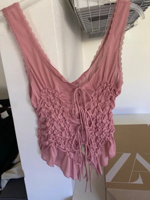 Rosa linnetopp med snörning och volang - Supersöt rosa linnetopp med snörning framtill och volangkant nedtill. Toppen har spetsdetaljer längs axelbanden och ett rynkat, nästan blommigt mönster över bröstet. Perfekt för dig som gillar romantisk och trendig stil.