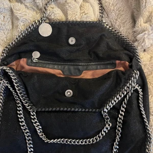 Svart Stella McCartney Falabella väska - Säljer en svart Falabella handväska från Stella McCartney med kedjedetaljer i silver runt kanterna och som axelrem. Väskan har magnetlås, innerfack med dragkedja och en rund logotypbricka. Tillverkad i veganskt läder med glansig finish. Små defekter på insidan.