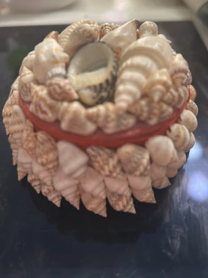 Snäckskalsprydd smyckesask - Unik rund smyckesask dekorerad med små snäckskal i olika former och färger. Asken är klädd i röd textil på insidan och har ett lock som också är täckt av snäckor. Perfekt för att förvara ringar eller små smycken.