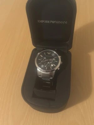 Emporio Armani herrklocka i stål - Snygg herrklocka från Emporio Armani med svart urtavla och silverfärgade detaljer. Klockan har kronograf och stilren länk i rostfritt stål. Levereras i originalbox med logga. Perfekt accessoar för en modern look.