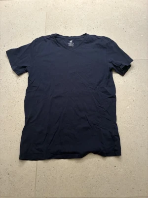 Mörkblå V-ringad T-shirt H&M - En enkel och stilren mörkblå T-shirt från H&M med V-ringning. Tillverkad i mjuk ekologisk bomull som känns skön mot huden. Perfekt basplagg för alla tillfällen och lätt att matcha med andra plagg.