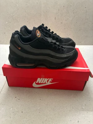 Nike Air Max 95 svarta sneakers - Nike Air Max 95 sneakers i svart med grå detaljer och orange logga på plösen. Skorna har en chunky sula med synlig Air-enhet och är tillverkade i en mix av mesh och mocka. Perfekta för dig som gillar streetwear och sportig stil.