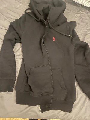 Svart loungewear-set från Ralph Lauren - Mysigt svart loungewear-set från Ralph Lauren med hoodie och matchande byxor. Hoodien har dragkedja, fickor och dragsko i huvan samt röd logga på bröstet. Byxorna har resår i midjan, dragsko och röd logga på benet. Perfekt för chill dagar hemma. Byxorna är M och tröjan S men byxorna sitter som S jag är 178 cm. Hör av dig vid minst fundering😄