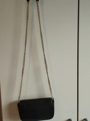 Svart axelväska med kedjerem - Snygg svart axelväska i slätt konstläder med lång kedjerem i metall. Väskan är ganska liten så har man en liten mobil får den plats, eller perfekt om man ger den till sina barn eller yngre personer 
