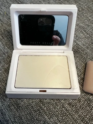 Fenty Beauty highlighter i puderform - Highlighter från Fenty Beauty i puderform med spegel i locket. Kommer i en vit fyrkantig dosa och har en ljus, champagnefärgad ton som ger glow till huden. Dosan har en beige insida med Fenty Beauty-logga. Perfekt för att framhäva ansiktets drag.