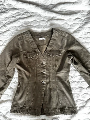 Olivgrön figursydd jeansjacka - Snygg olivgrön jeansjacka från Zara Basic med figurnära passform, V-ringning och fyra knappar framtill. Jackan har två bröstfickor med lock och dekorativa sömmar som ger en cool siluett. Perfekt för dig som vill ha en unik twist på klassiska jeansjackan.