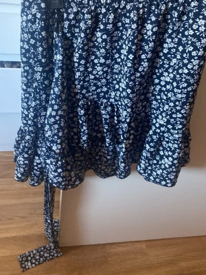 Blommig kortkjol med volang - Söt kortkjol med mörkblå botten och vita blommor. Kjolen har volang nertill och knytband i sidan som ger en omlott-look. Perfekt för dig som gillar romantiska och trendiga plagg. Materialet känns lätt och luftigt.