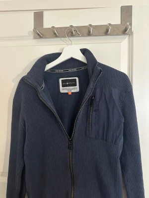 Blå ziptröja från Sail Racing - Mörkblå ribbstickad ziptröja från Sail Racing med hög krage och dragkedja framtill. Bröstficka med dragkedja och logga på vänster sida. Tröjan har en sportig look och är tillverkad i ett mjukt material som känns skönt mot huden.