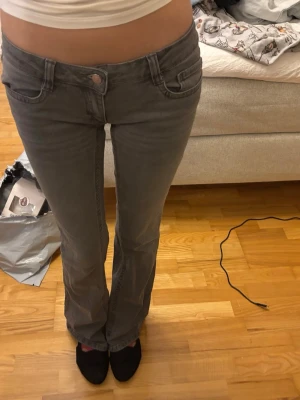 Grå low waisted bootcut jeans från Din Vintage Shop - Säljer ett par gråa low waisted bootcut jeans från Din Vintage Shop. Jeansen har klassisk femficksdesign, dekorativa sömmar på bakfickorna och knappgylf. Materialet är mjukt denim och passformen är något utsvängd nedtill. Perfekta för en avslappnad och trendig look.