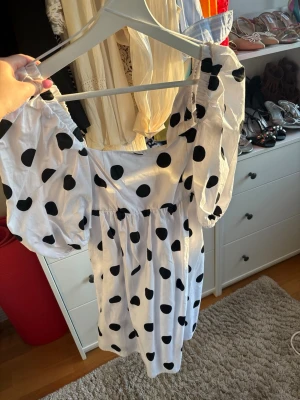 Vit kort klänning med svarta prickar polka - Supersöt vit kortklänning med stora svarta prickar och puffiga kortärmar. Klänningen har en markerad midja och en lätt rynkad kjol. Materialet känns lätt och luftigt, perfekt för varma dagar. Snygg urringning och elastiska detaljer vid axlarna.