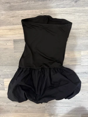 Svart axelbandslös kort klänning - Säljer en svart axelbandslös kort klänning med puffig kjol. Klänningen har en ballongliknande nederdel och är perfekt för dig som vill sticka ut. Materialet känns som polyester och modellen är riktigt trendig. (Egna bilder) skriv till mig vid frågor💕💕