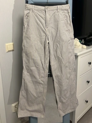 Randiga cargobyxor i vitt och blått - Säljer ett par cargobyxor i jeansmaterial med vita och blåa vertikala ränder. Byxorna har flera fickor på sidorna och baktill, samt raka ben och hällor för skärp. Perfekta för en avslappnad och stil. BARNSTORLEK 158!