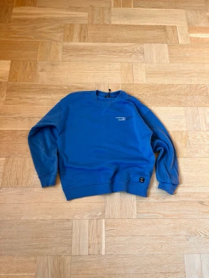 Blå sweatshirt från Björn Borg - Säljer en blå sweatshirt från Björn Borg med rund halsringning och ribbade muddar vid ärmar och nederkant. Tröjan har diskret logga på bröstet och en liten patch nertill. Perfekt för en avslappnad stil.