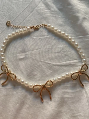 Pärlhalsband med rosetter Montini - Sött halsband från Montini med vita pärlor och tre guldfärgade rosetter som detaljer. Halsbandet har justerbar kedja och stängs med ett klassiskt lås. Perfekt accessoar för att ge din outfit en gullig och trendig touch.