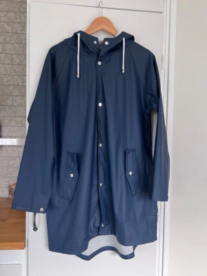 Mörkblå regnjacka med huva, storlek M - Marinblå regnjacka med huva, vita snören och två fickor. Tryckknappar framtill och en längre passform. Finns lite märken/fläckar som inte gått bort i tvätten.
