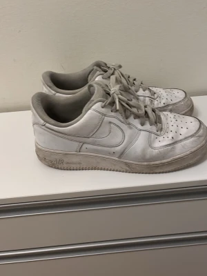 Nike Air Force 1 vita sneakers - Klassiska vita Nike Air Force 1 sneakers i lågt utförande. Skorna har den ikoniska swoosh-loggan på sidan, perforerad tåbox och AIR-detalj på sulan. Tillverkade i läder med textilfoder och snörning framtill. Perfekta för en clean och tidlös stil.
