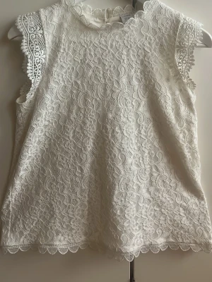 Vit spetsblus från Lindex - Elegant och vacker benvit blus från Lindex i spets med blommönster och scallopkant vid hals och ärmhål. Ärmlös modell med knappstängning bak i nacken. Perfekt för dig som gillar en feminin och söt look. Materialet är genombruten spets som ger en lyxig känsla. Endast använd 1 gång. Som ny! Storleken är stor för att vara 170.